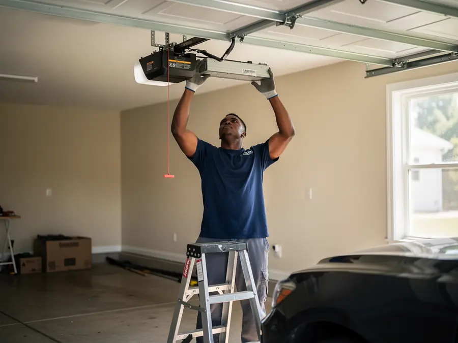 A technician on a ladder installs a garage door opener in a garage.