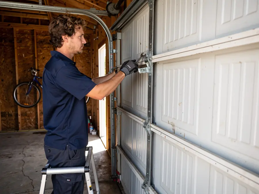 A technician on a ladder adjusts a white garage door.